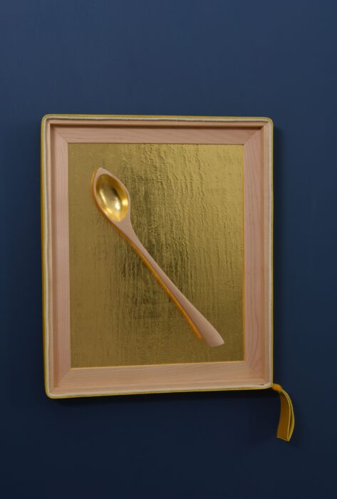 Hand-carved birch spoon resting against a textured gold leaf background, framed in birch with leather and felt accents.